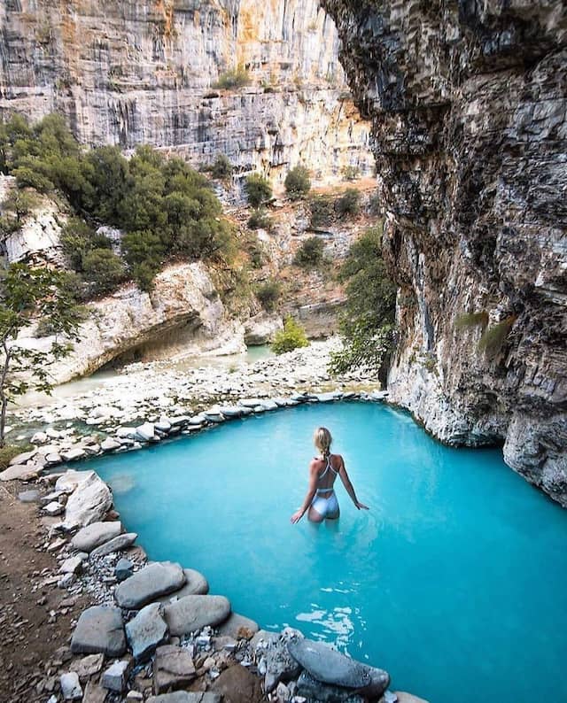 Woman in white bikini walking in thermal bath in benja filled with blue water and surrounded by rocks