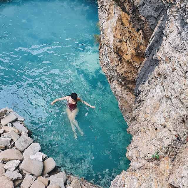 Woman in brown bikini swimming in thermal bath in benja filled with blue water and surrounded by rocks