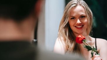 Blonde smiling girl flirting with a man on table while holding a red rose