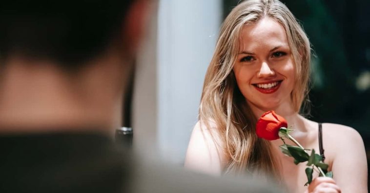 Blonde smiling girl flirting with a man on table while holding a red rose