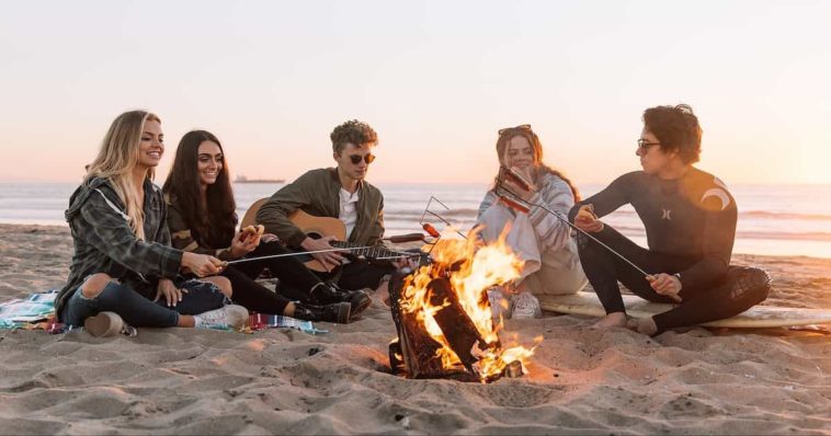 Five young people sitting around a campfire at the beach during sunset playing acoustic guitar and eating sausages