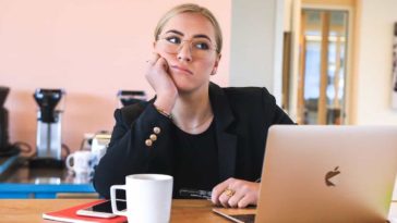 Sad blonde woman dressed in black is sitting in front of a silver macbook air