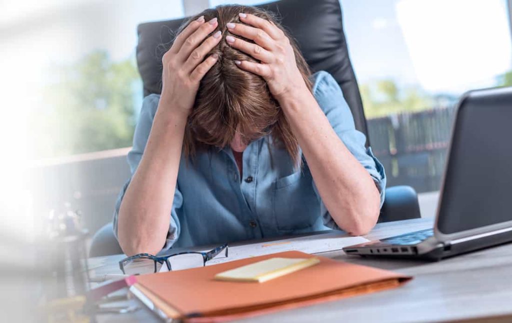 stressed and tired woman in blue shirt sitting on her desk with her hands on her head