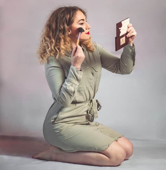 A woman dressed in a green coat putting makeup while holding a small mirror