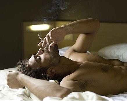 A naked man smoking cigarrete while lying on his bed
