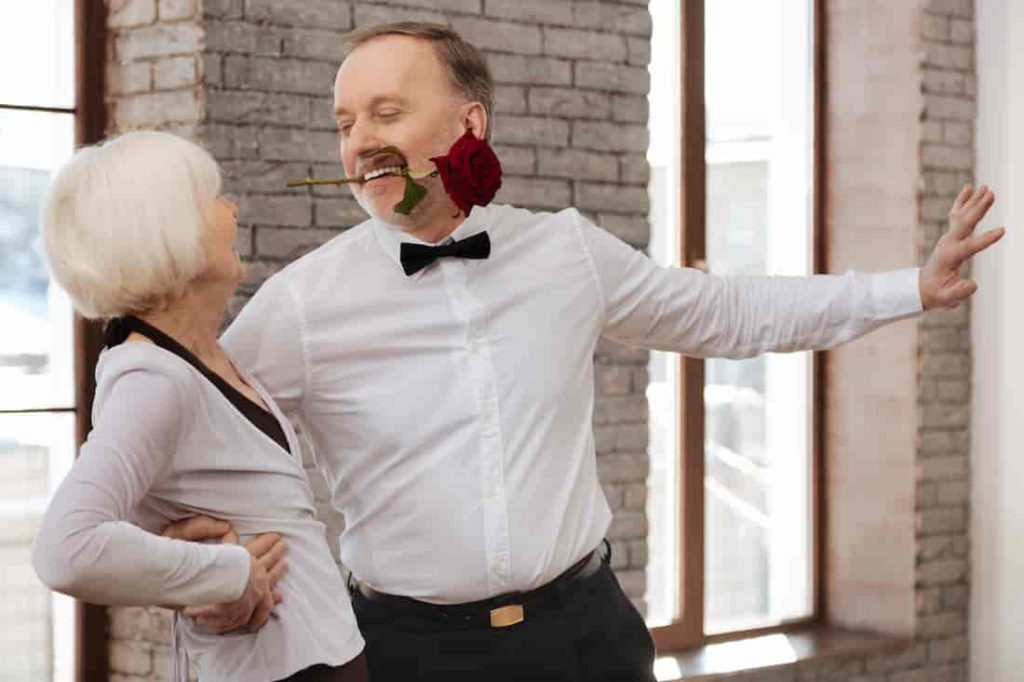 man in a white shirt with a red rose in his mouth dances with a white haired woman